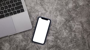A phone with a blank white screen lying on concrete next to the corner of a laptop