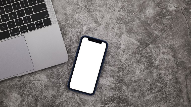 A phone with a blank white screen lying on concrete next to the corner of a laptop