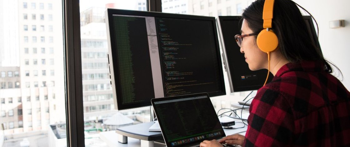 A developer in headphones at a two-monitor desk, code open on both screens, city windows behind