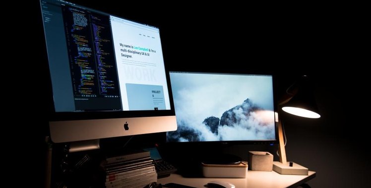 A dark desk lit by a single lamp, two monitors showing a web page and a photo wallpaper