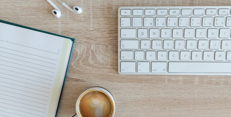 A desk seen from above: a blank notebook, a white keyboard, earbuds and a cup of coffee on light wood