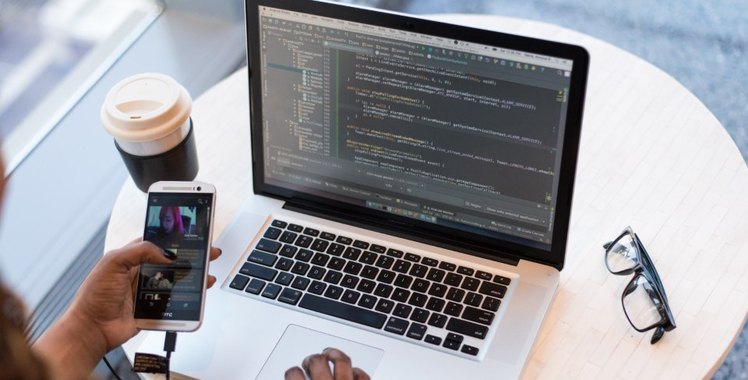 Hands on a laptop keyboard with code on the screen, a phone held in one hand, a coffee cup beside it